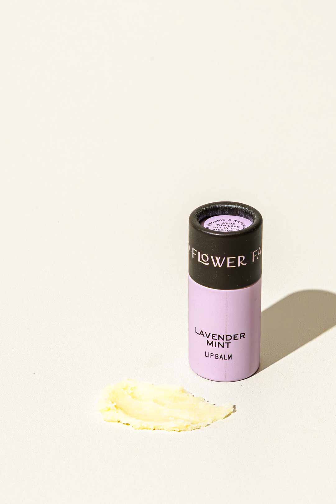 A lavender-colored biodegradable tube labeled Lavender Mint Organic Lip Balm from Good Flower Farm stands upright on a light surface with a small smear of balm beside it. The cap reads FLOWER FAR.