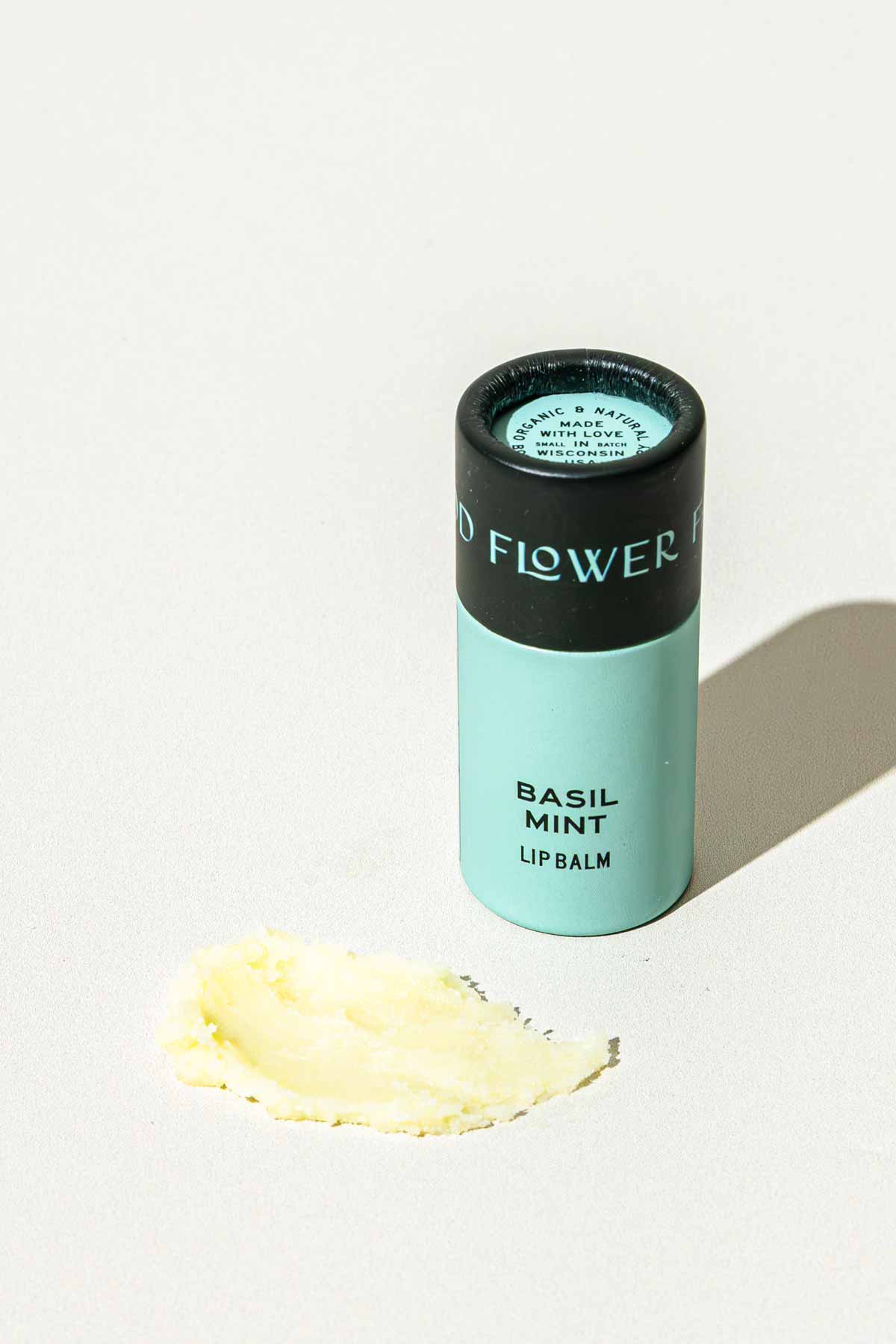 A light blue tube of Good Flower Farm’s Basil Mint Lip Organic Lip Balm stands upright on a white surface, with a smear of the balm spread out beside it.
