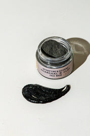 An open jar of Good Flower Farm’s TESTER — Charcoal & Clay Botanical Face Mask sits on a white surface, a dark smear in front. The label reads “Detoxifying & Clearing Charcoal Clay Face Mask.”.