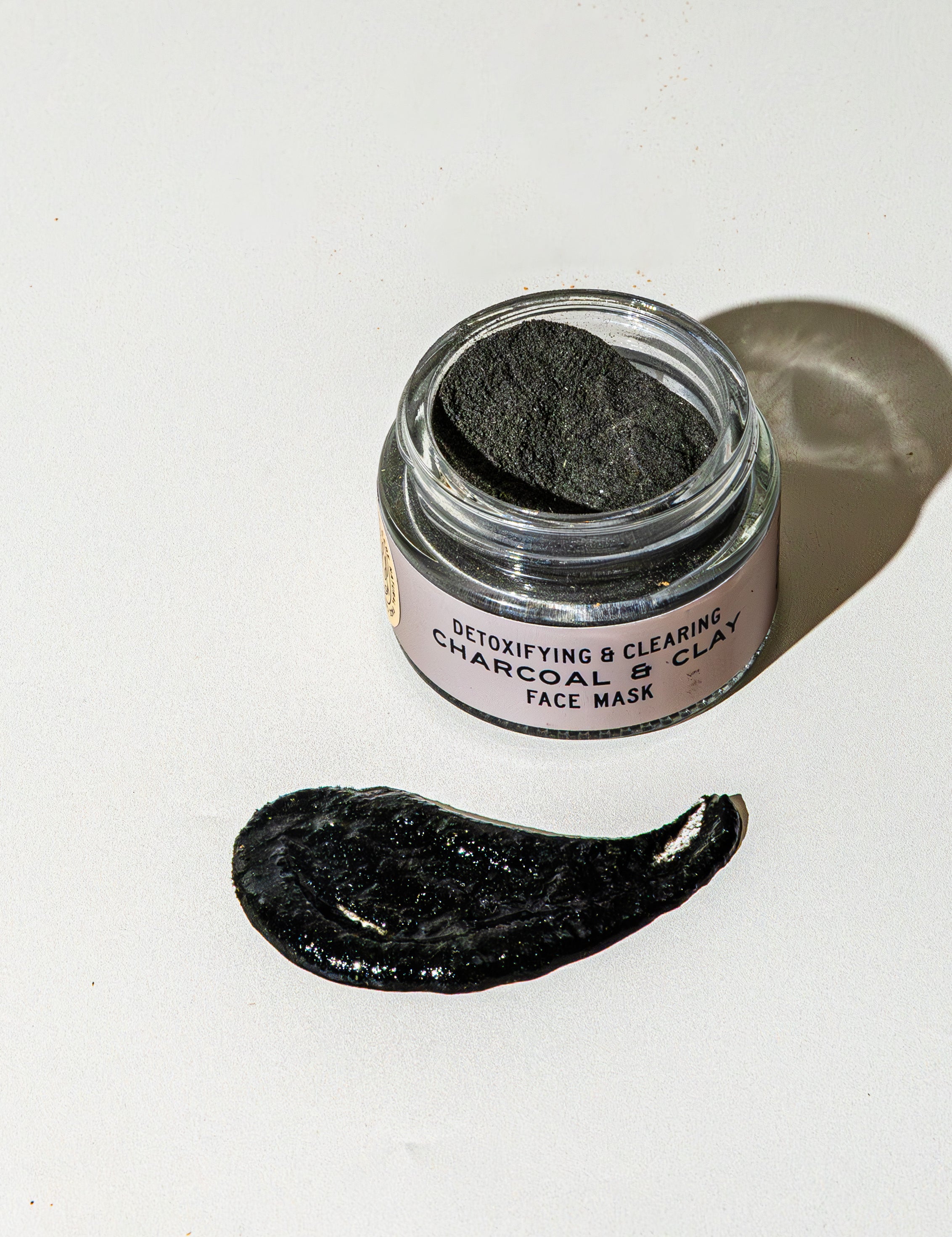 An open jar of Good Flower Farm’s TESTER — Charcoal & Clay Botanical Face Mask sits on a white surface, a dark smear in front. The label reads “Detoxifying & Clearing Charcoal Clay Face Mask.”.