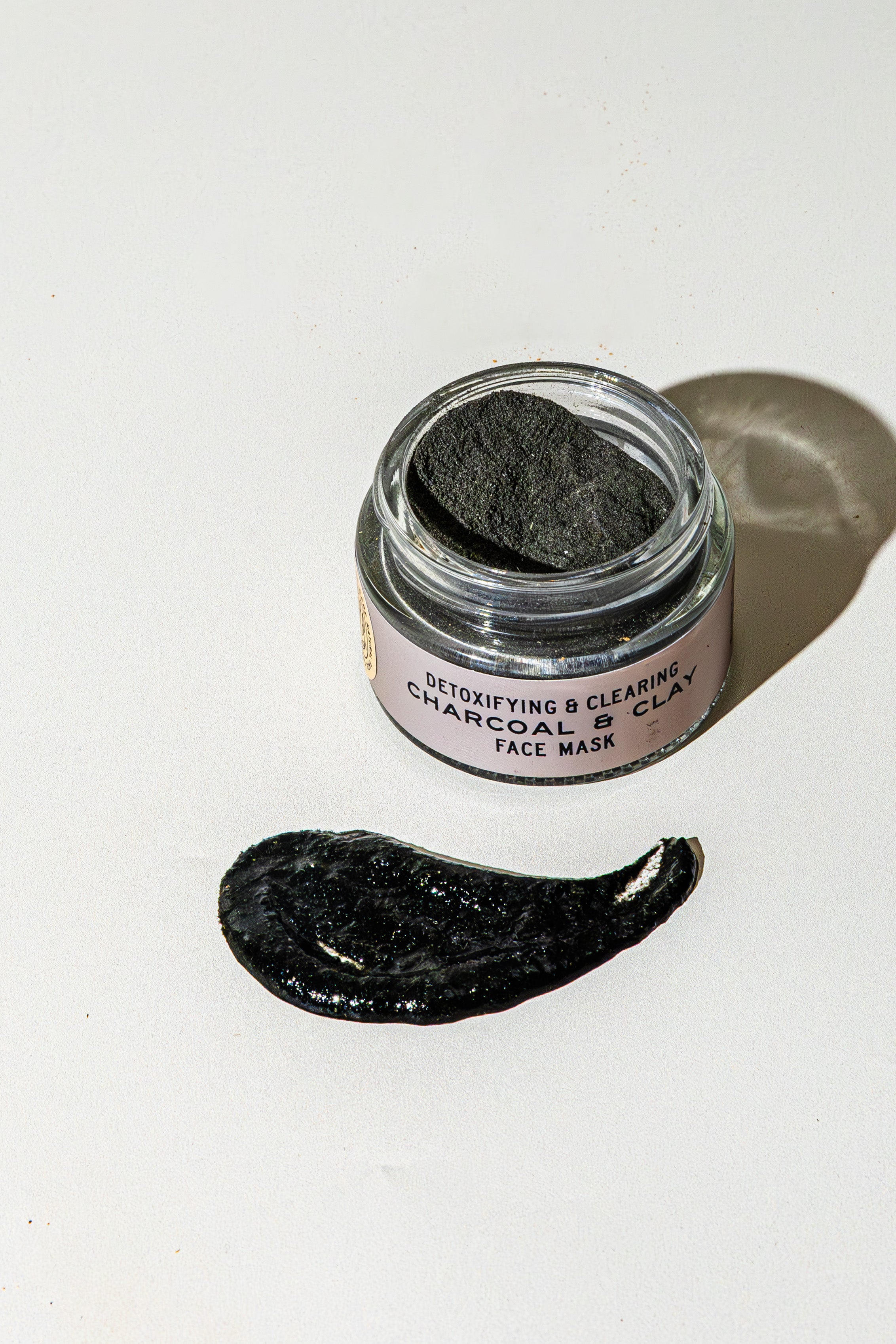 An open jar of Good Flower Farm’s TESTER — Charcoal & Clay Botanical Face Mask sits on a white surface, a dark smear in front. The label reads “Detoxifying & Clearing Charcoal Clay Face Mask.”.