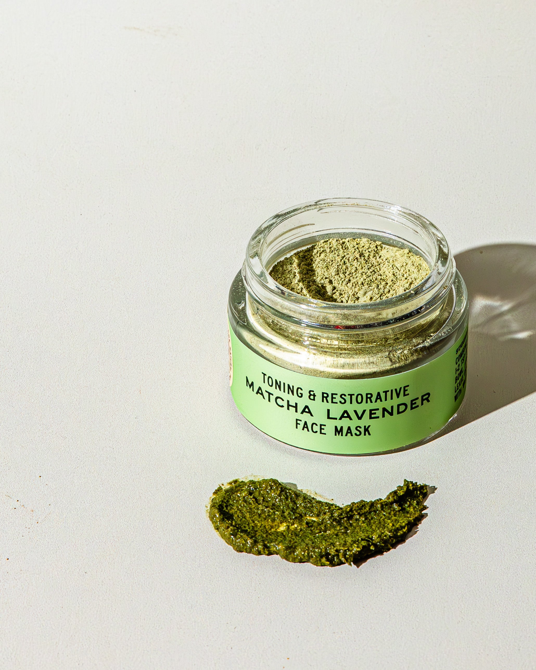 An open glass jar labeled TESTER — Matcha Lavender Botanical Face Mask by Good Flower Farm sits on a white surface, with a smear of green matcha powder and French green clay spread nearby.