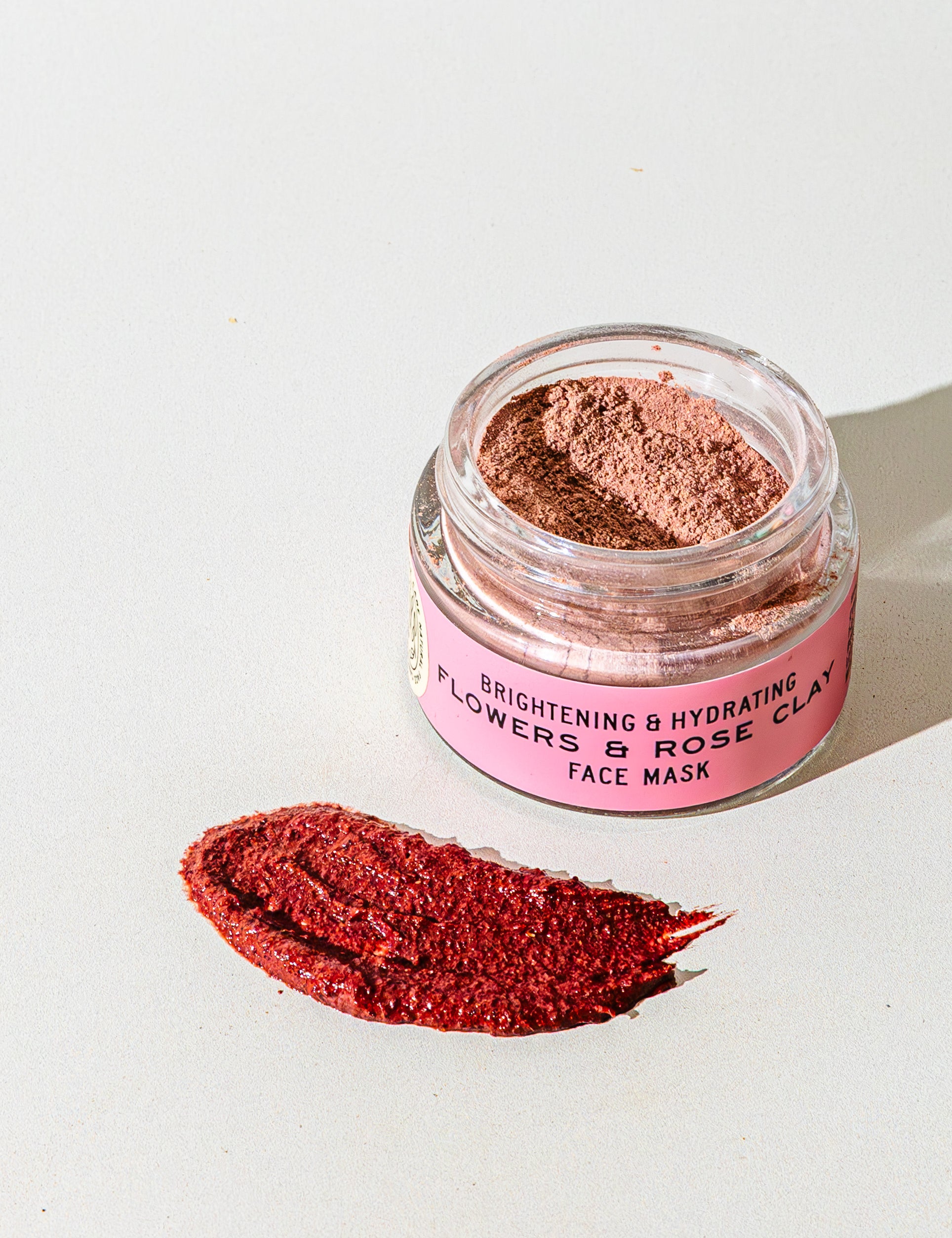 An open jar of TESTER — Flowers & Rose Clay Botanical Face Mask by Good Flower Farm rests on a light surface, showcasing its botanical formula with a swatch of the rosy clay mask displayed in front.