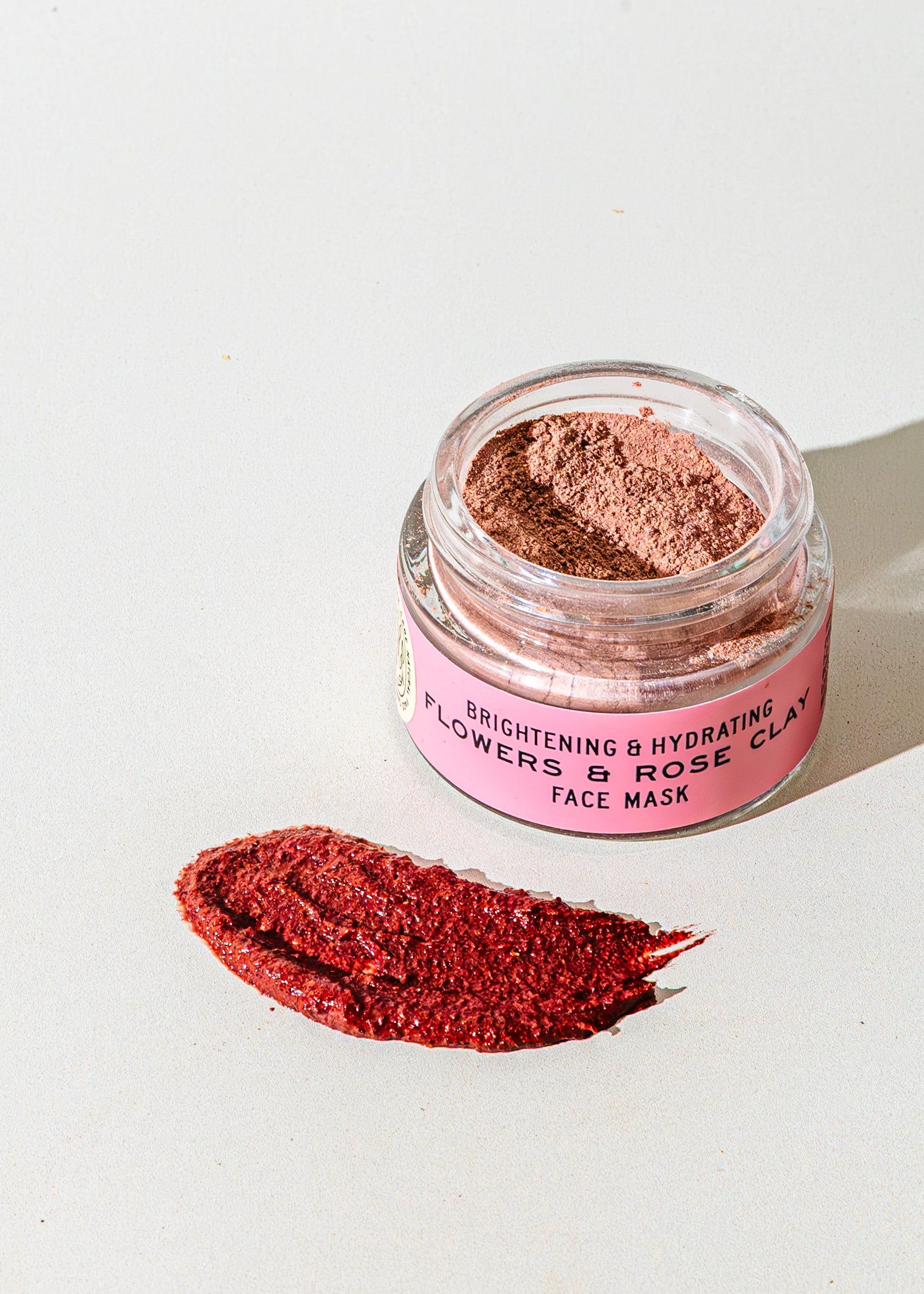 An open jar of TESTER — Flowers & Rose Clay Botanical Face Mask by Good Flower Farm rests on a light surface, showcasing its botanical formula with a swatch of the rosy clay mask displayed in front.