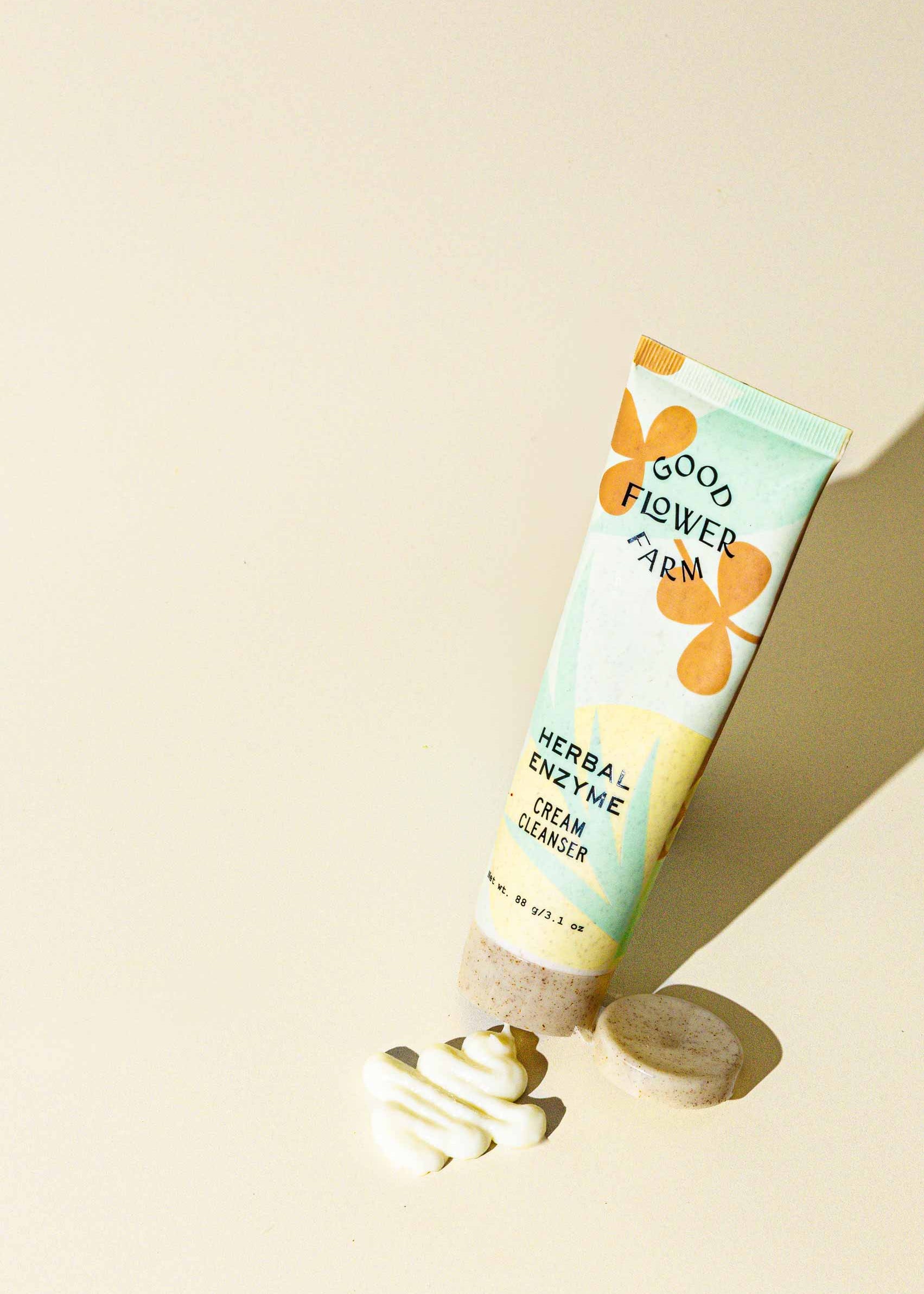A tube of Good Flower Farm Herbal Enzyme Cream Cleanser & Makeup Remover stands upright on a light surface beside a round bar and a swirl of white cream, highlighted by bright natural lighting.