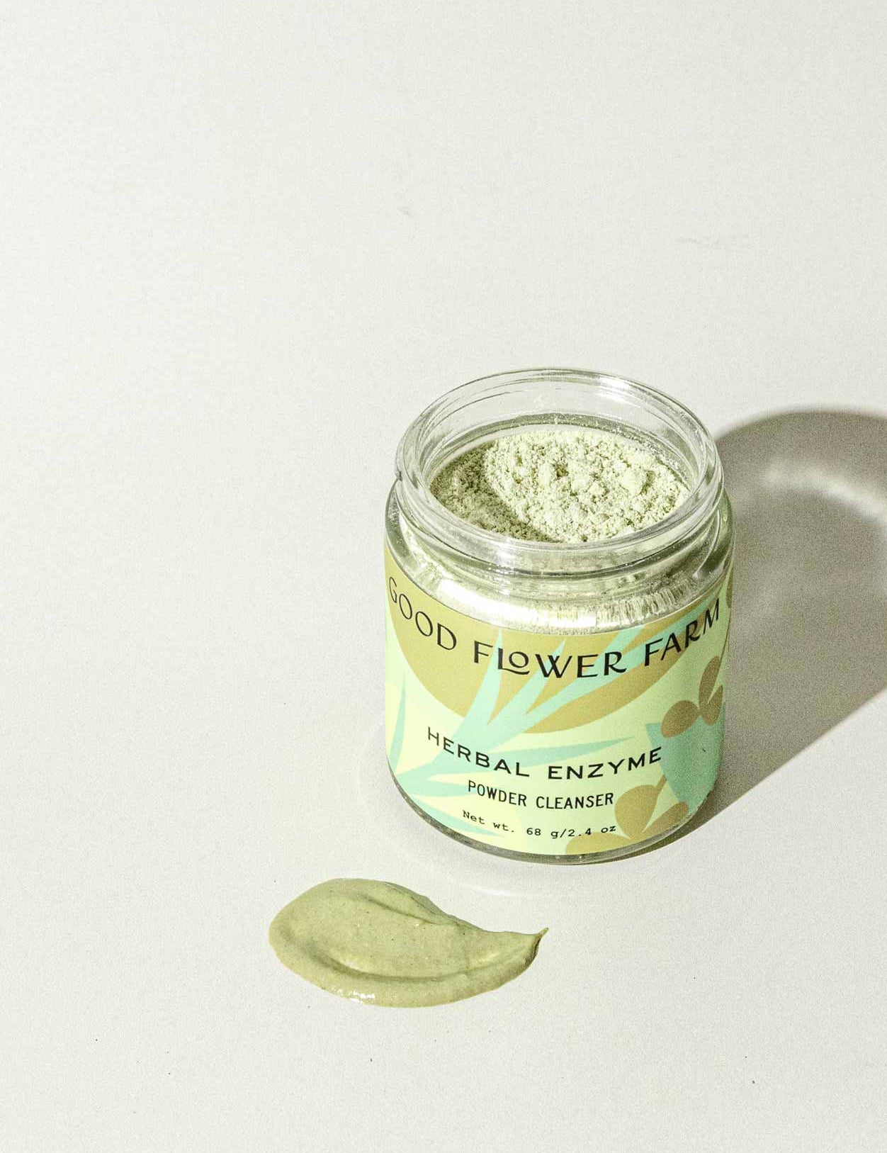 A jar of Good Flower Farm Herbal Enzyme Powder Facial Cleanser sits on a light surface, with some green powder—featuring papaya and pineapple enzymes—spread out in front of it.