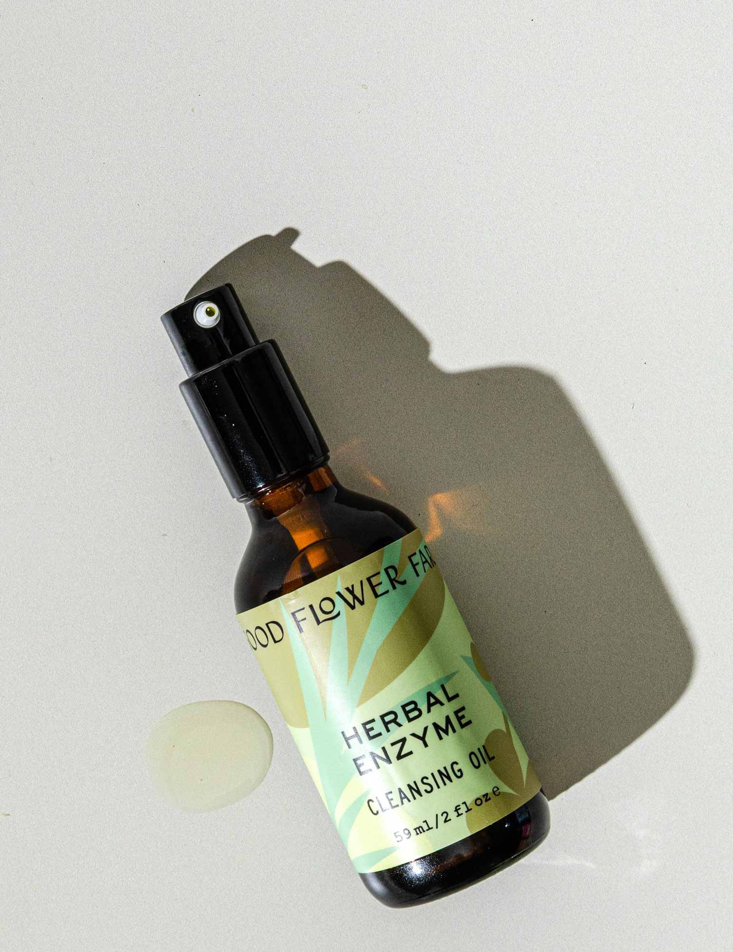 A brown glass spray bottle of Good Flower Farms Herbal Enzyme Cleansing Oil, featuring a green label with leaf graphics, rests on a light surface beside a small oil spill and casts a shadow, highlighting its purifying, papaya seed oil blend.
