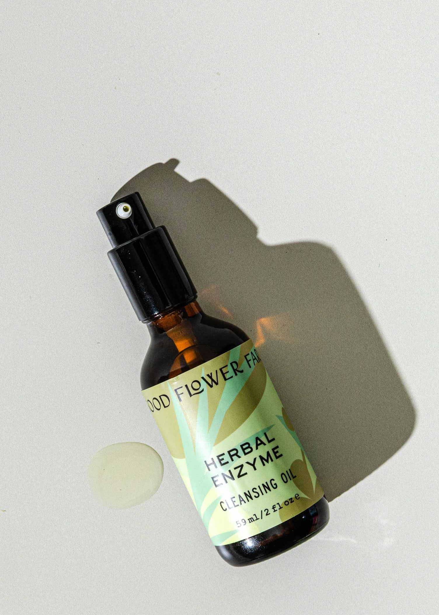 A brown glass spray bottle of Good Flower Farms Herbal Enzyme Cleansing Oil, featuring a green label with leaf graphics, rests on a light surface beside a small oil spill and casts a shadow, highlighting its purifying, papaya seed oil blend.
