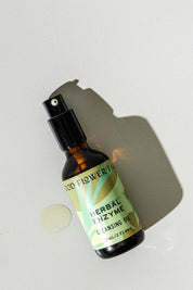 A brown glass spray bottle of Good Flower Farms Herbal Enzyme Cleansing Oil, featuring a green label with leaf graphics, rests on a light surface beside a small oil spill and casts a shadow, highlighting its purifying, papaya seed oil blend.