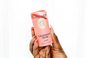 A hand with long, pink nails holds a cylindrical pink container labeled Good Flower Farm Calendula Rose Salve—an herbal, eco-friendly salve—against a plain white background.
