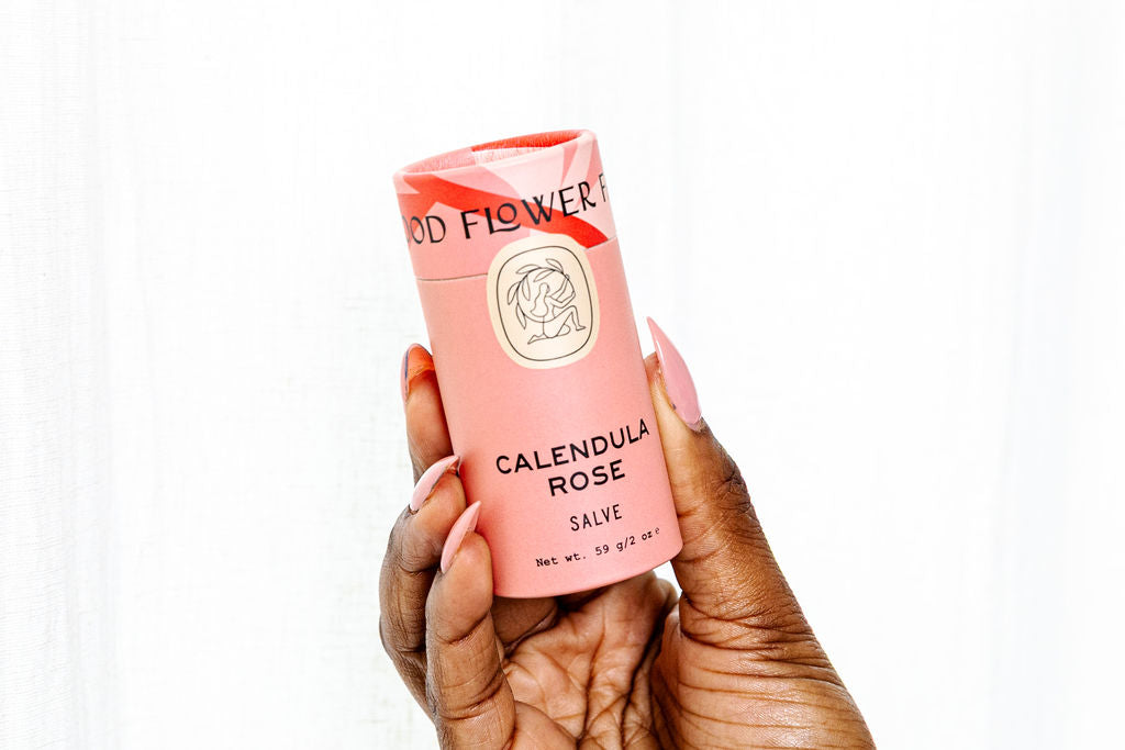 A hand with long, pink nails holds a cylindrical pink container labeled Good Flower Farm Calendula Rose Salve—an herbal, eco-friendly salve—against a plain white background.