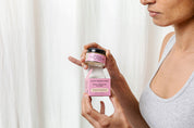A person in a gray tank top opens a jar of Good Flower Farm Rose Geranium Face Cream with evening primrose oil, holding it and the matching pink box so both labels are visible against a light background.