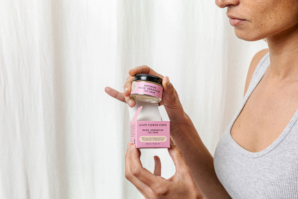 A person in a gray tank top opens a jar of Good Flower Farm Rose Geranium Face Cream with evening primrose oil, holding it and the matching pink box so both labels are visible against a light background.