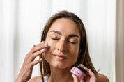 A woman with closed eyes applies Good Flower Farm Rose Geranium Face Cream to her cheek, holding a small jar with a pink label in front of a light curtain.
