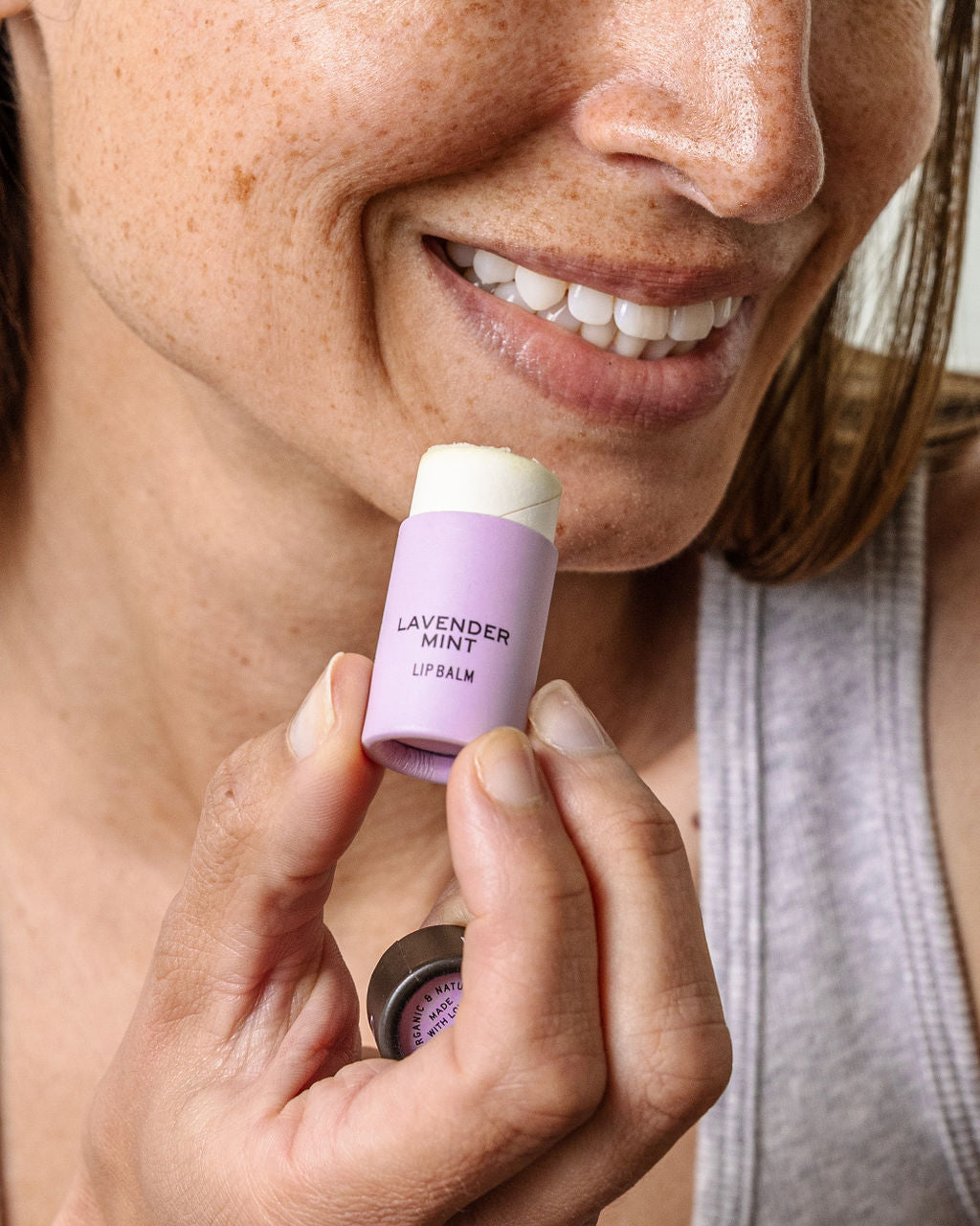 A smiling person holds a tube of Good Flower Farm’s Lavender Mint Organic Lip Balm close to their lips, the biodegradable tubes label clearly visible as they prepare to apply it.