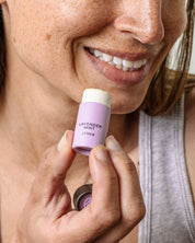 A smiling person holds a tube of Good Flower Farm’s Lavender Mint Organic Lip Balm close to their lips, the biodegradable tubes label clearly visible as they prepare to apply it.