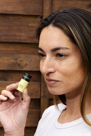 A woman with light brown hair and freckles holds a Good Flower Farm Lemon Vanilla Organic Lip Balm near her face, standing in front of a wooden background.