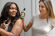 Two women smile as they hold bottles of Good Flower Farms Into the Woods Natural Bugspray toward each other, standing in front of a light gray wooden wall. One wears a black tank top with long dark hair; the other, a white t-shirt and light brown hair.