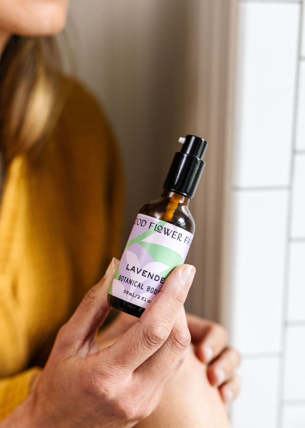 A person in a mustard yellow top holds a small bottle labeled “Lavender Botanical Body Oil” by Good Flower Farm, featuring a pastel design, in front of a white tiled wall.