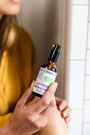 A person in a mustard yellow top holds a small bottle labeled “Lavender Botanical Body Oil” by Good Flower Farm, featuring a pastel design, in front of a white tiled wall.