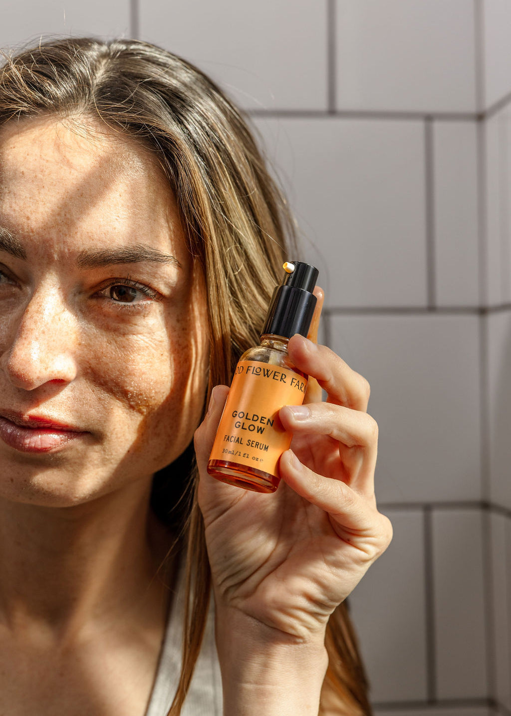 A woman with long brown hair holds up a bottle of Good Flower Farms Golden Glow Facial Serum in sunlight against a tiled wall, showcasing the serums promise of natural radiance and skin hydration.