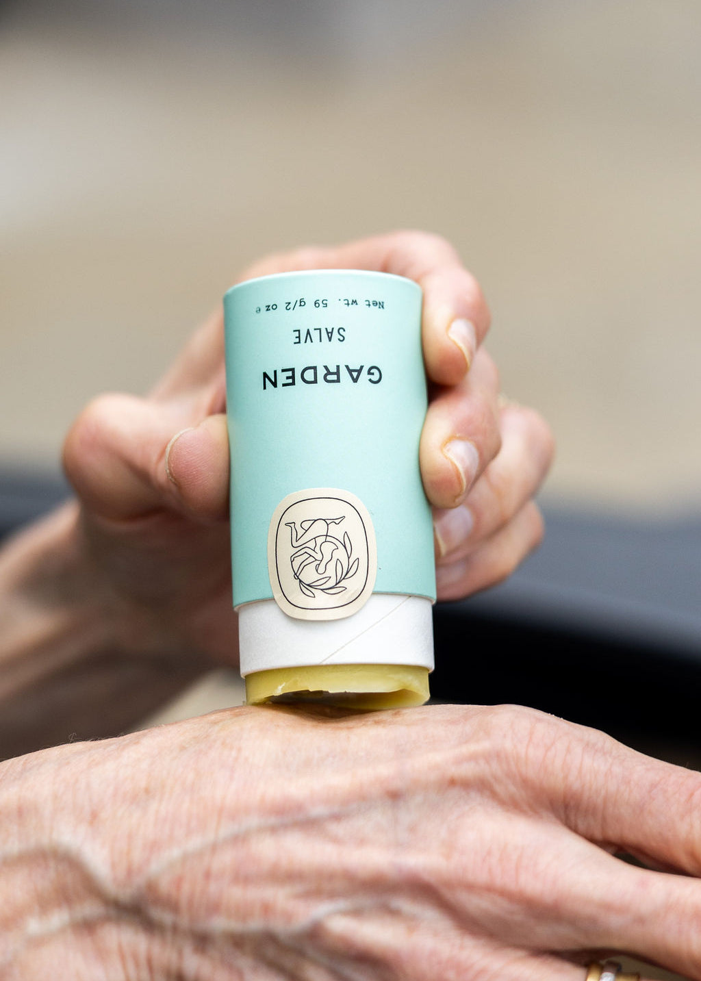 A person applies Good Flower Farms Garden Salve from a light blue tube with a beige cap and botanical logo to their hand; the salve is crafted with nourishing farm-grown herbs to soothe skin.