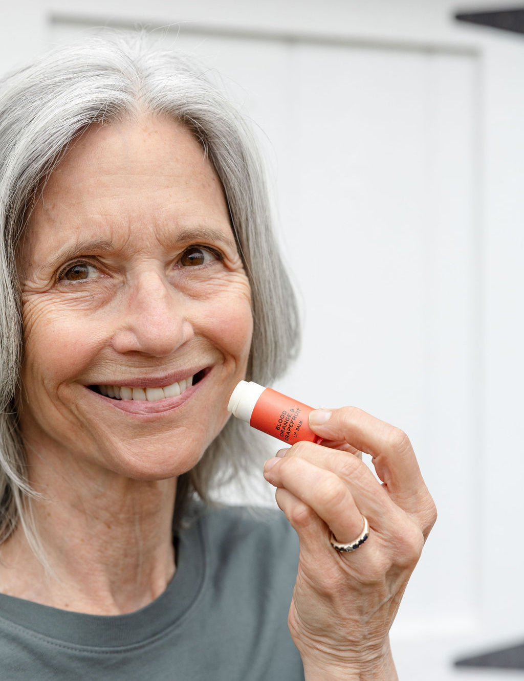 An older woman with gray hair smiles while holding Good Flower Farms Blood Orange & Grapefruit Organic Lip Balm near her lips, standing in front of a white background and wearing a gray shirt.