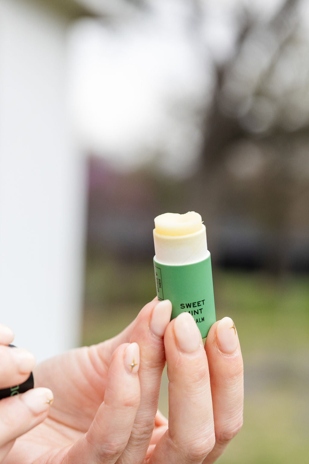 With manicured nails, someone holds an open tube of Good Flower Farm Sweet Mint Organic Lip Balm outdoors, with trees and grass softly blurred in the background.