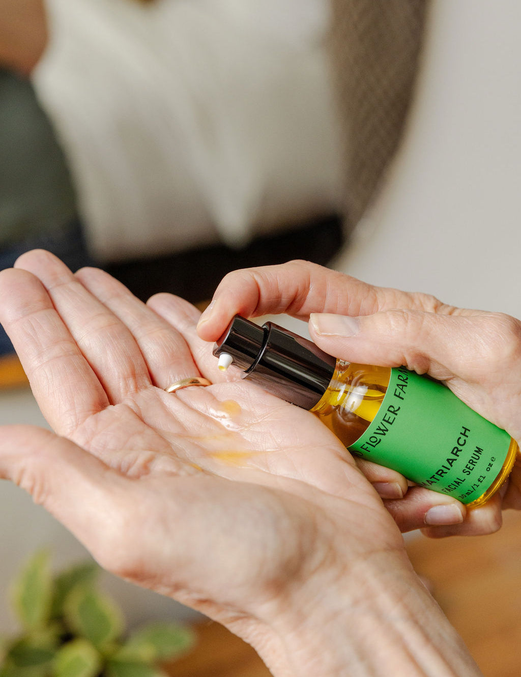 A person dispenses a few drops of Matriarch Facial Serum by Good Flower Farm from a small green bottle into their open palm, with a blurred background and a small plant in view.