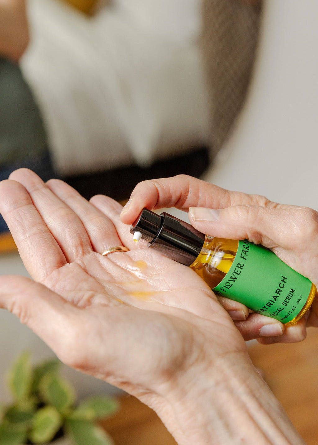 A person dispenses a few drops of Matriarch Facial Serum by Good Flower Farm from a small green bottle into their open palm, with a blurred background and a small plant in view.