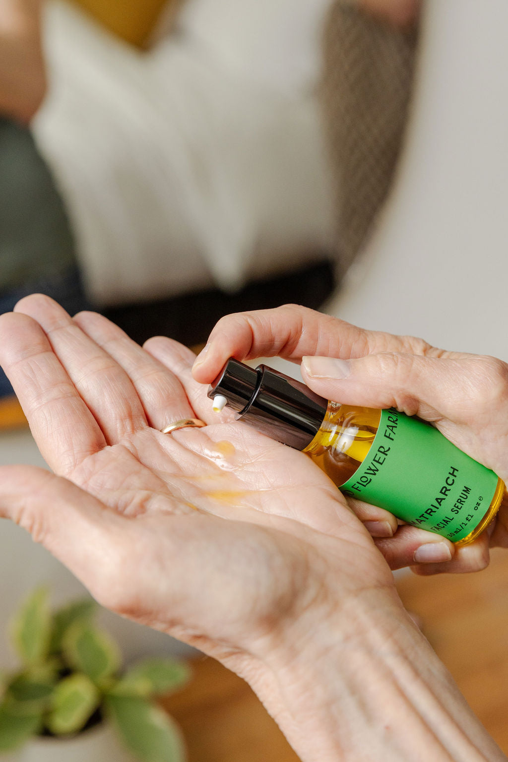 A person dispenses a few drops of Matriarch Facial Serum by Good Flower Farm from a small green bottle into their open palm, with a blurred background and a small plant in view.