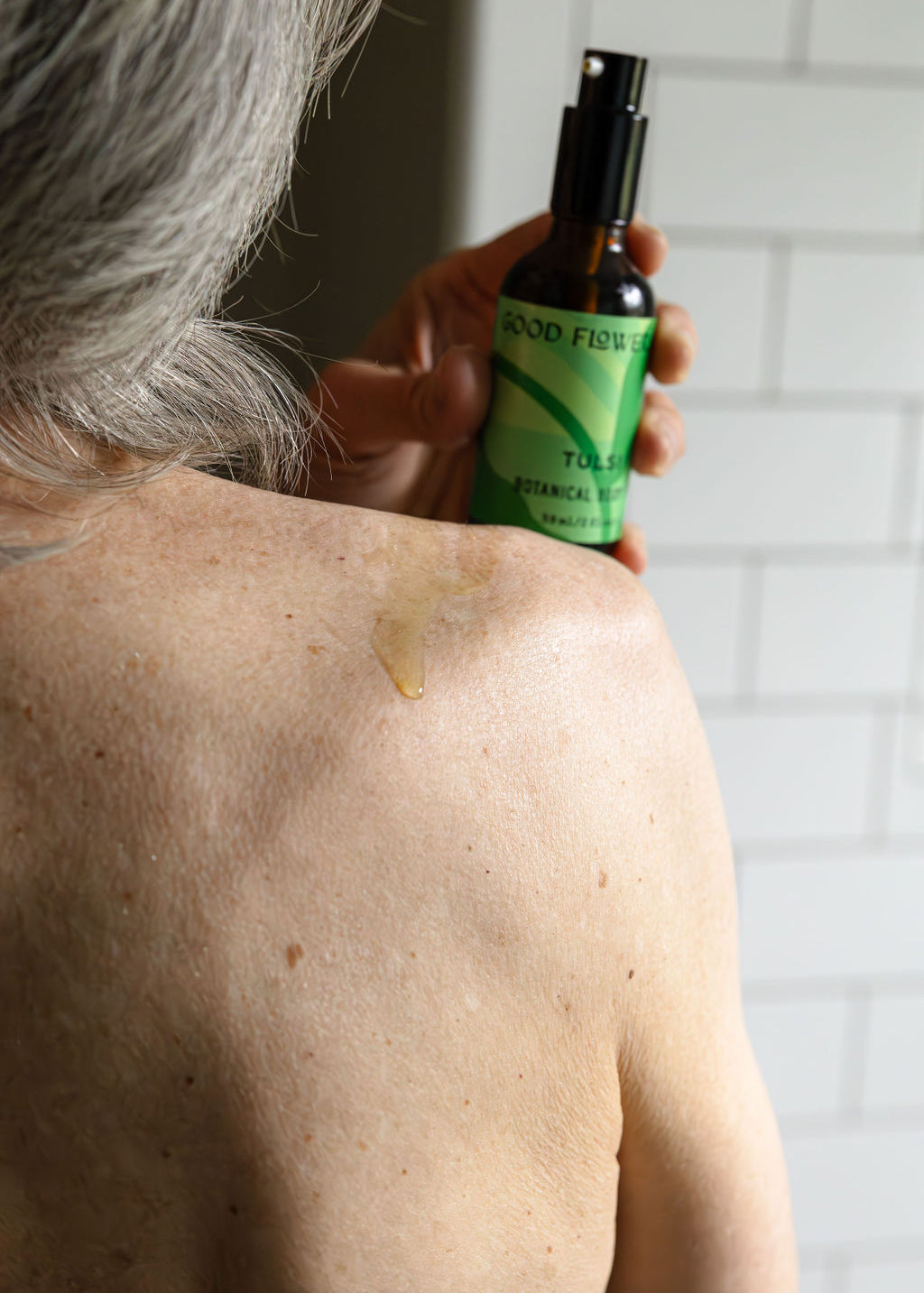 A person with gray hair applies Good Flower Farms Tulsi Botanical Body Oil from a green-labeled bottle onto their bare shoulder, a drop glistening on their skin against a white tiled wall backdrop.
