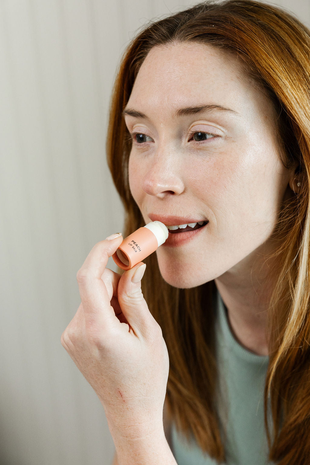 A fair-skinned, red-haired woman in a light green top applies Good Flower Farm’s Peach Organic Lip Balm, an eco-friendly, light pink balm, to her lips while gazing to the side against a pale background.