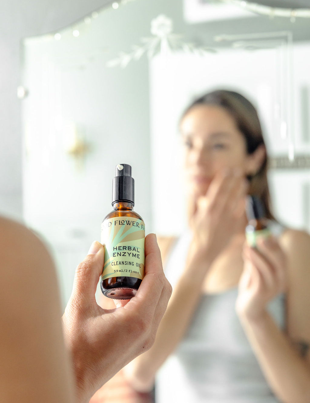 A person holds a bottle of Good Flower Farm Herbal Enzyme Cleansing Oil in focus, while their blurred reflection applies the product to their face in front of a mirror.