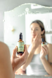 A person holds a bottle of Good Flower Farm Herbal Enzyme Cleansing Oil in focus, while their blurred reflection applies the product to their face in front of a mirror.