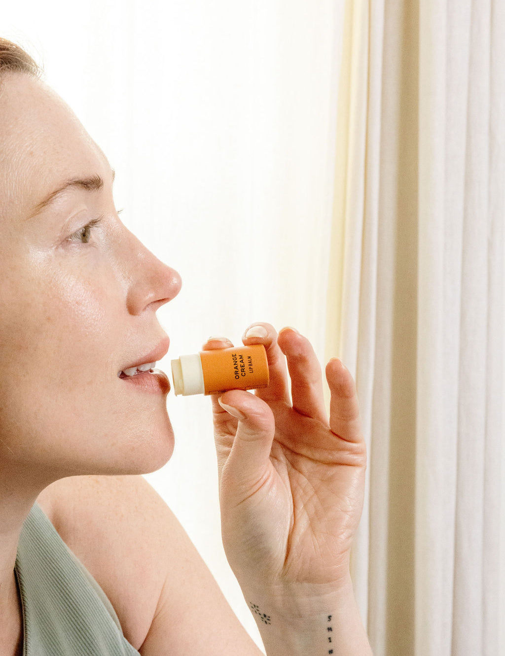 A woman with light skin, wearing a sleeveless top, holds a Good Flower Farm Orange Cream Organic Lip Balm near her lips in front of light-colored curtains, ready to apply the moisturizing balm.