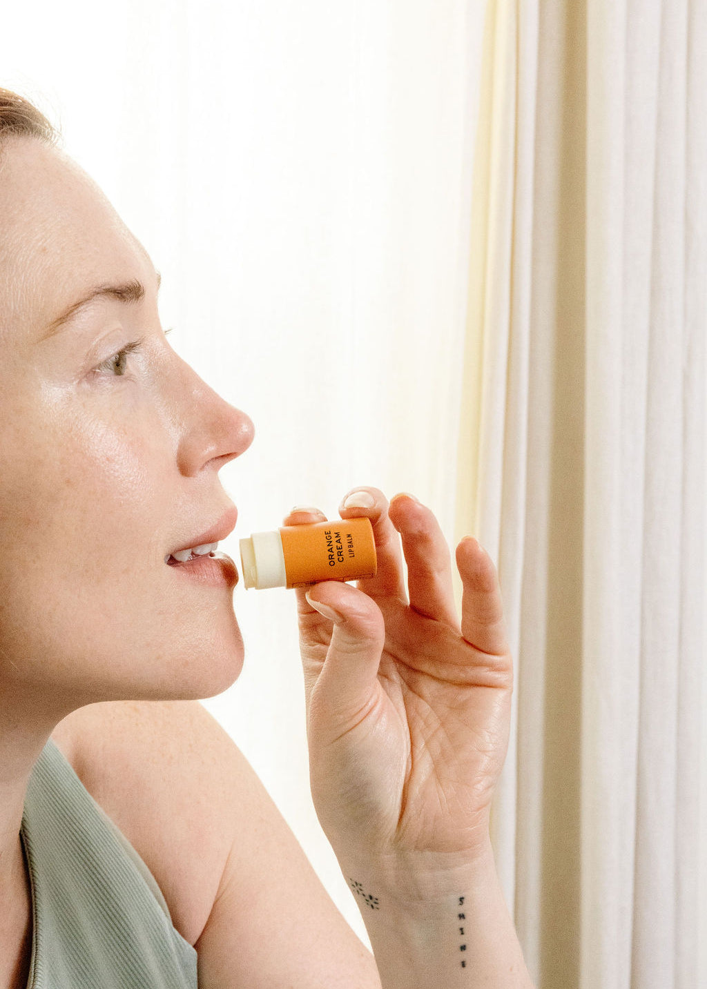 A woman with light skin, wearing a sleeveless top, holds a Good Flower Farm Orange Cream Organic Lip Balm near her lips in front of light-colored curtains, ready to apply the moisturizing balm.