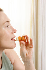 A woman with light skin, wearing a sleeveless top, holds a Good Flower Farm Orange Cream Organic Lip Balm near her lips in front of light-colored curtains, ready to apply the moisturizing balm.