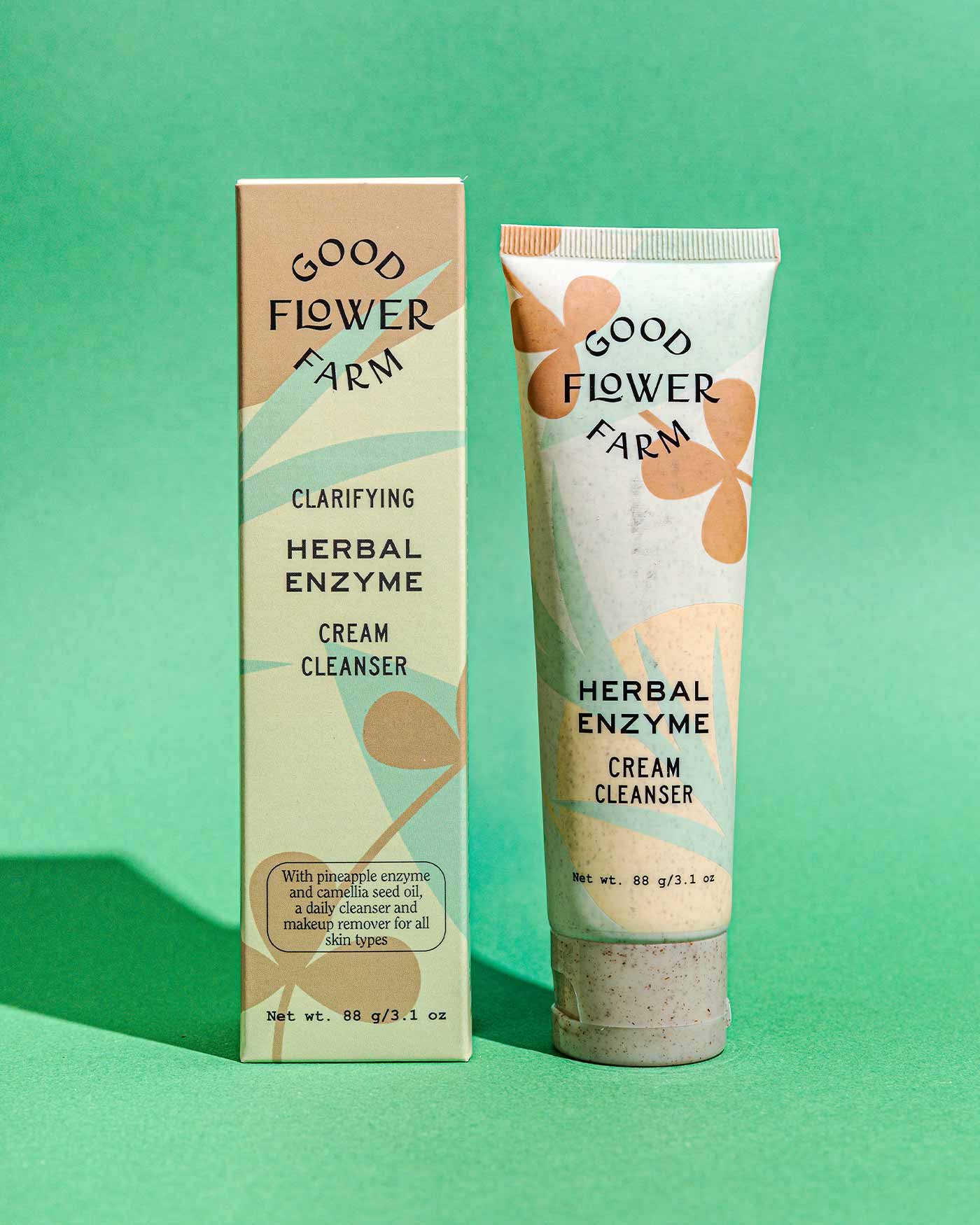 A tube and box of Good Flower Farm Herbal Enzyme Cream Cleanser & Makeup Remover, featuring abstract leaf designs and product details, are displayed on a green background.