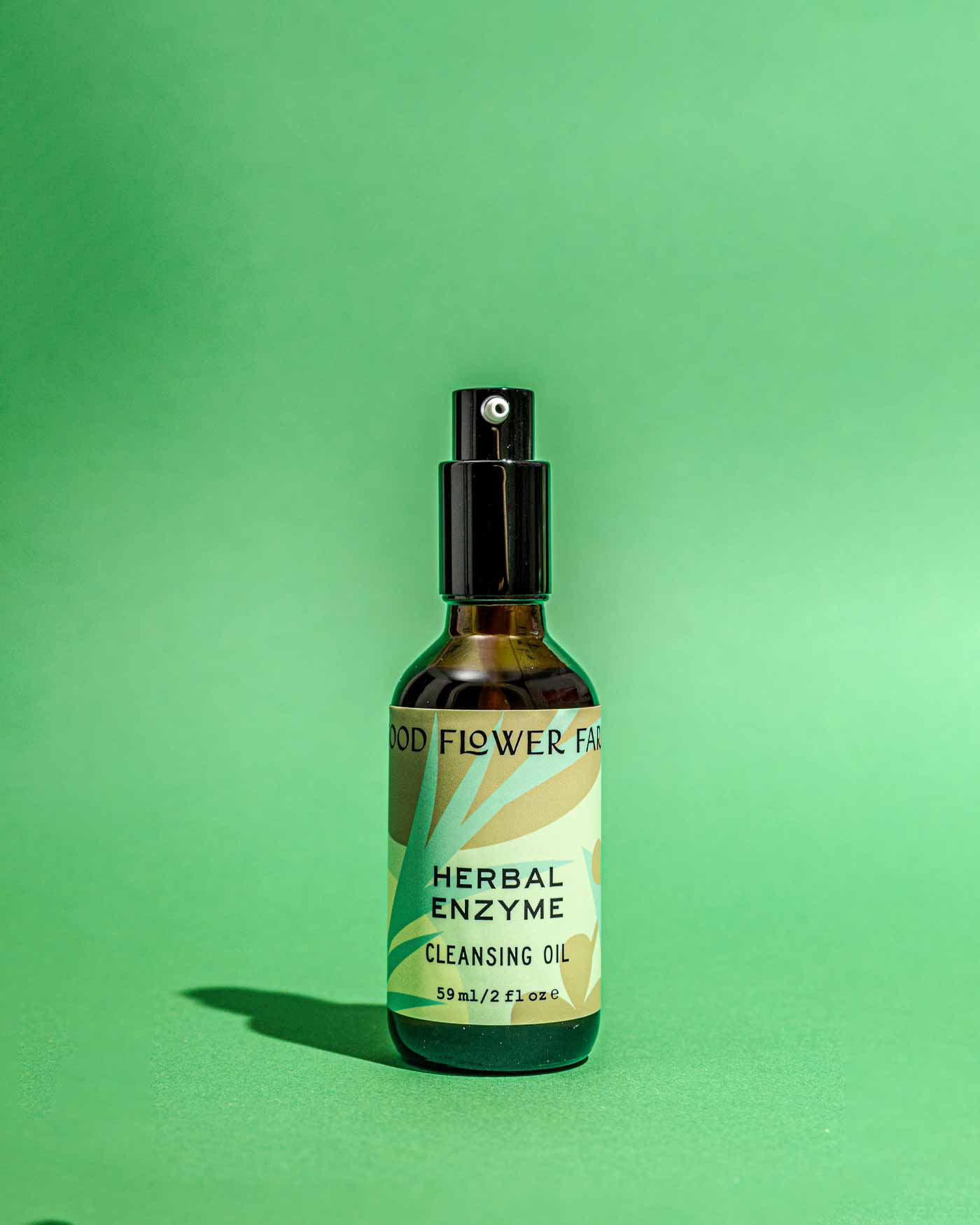 A bottle of Good Flower Farm Herbal Enzyme Cleansing Oil with a spray top sits on a green surface. The label, decorated with leaves and earthy tones, highlights this papaya seed oil-infused cleanser’s purifying oil-to-milk formula.