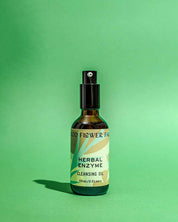 A bottle of Good Flower Farm Herbal Enzyme Cleansing Oil with a spray top sits on a green surface. The label, decorated with leaves and earthy tones, highlights this papaya seed oil-infused cleanser’s purifying oil-to-milk formula.