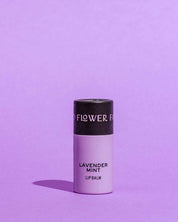 A lavender-colored biodegradable tube of Good Flower Farm’s Lavender Mint Organic Lip Balm stands upright on a matching lavender background, casting a left shadow. This soothing balm is enriched with nourishing coconut oil.