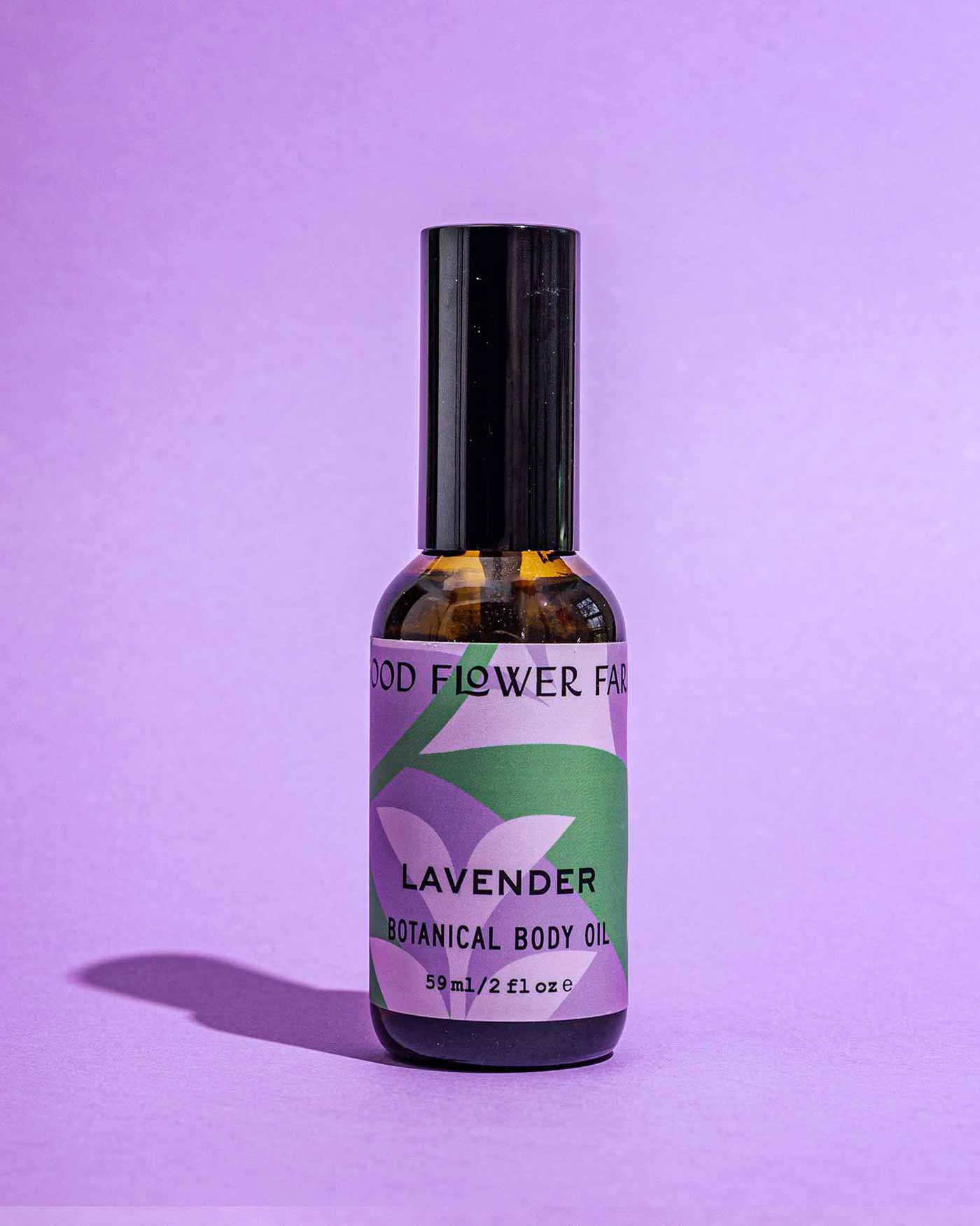 A bottle of Good Flower Farm’s TESTER — Lavender Botanical Body Oil stands upright on a purple surface with a matching background. The label, with green leaves and purple text, highlights nourishing lavender oil and hemp seed oil.