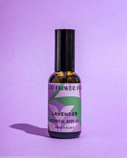 A bottle of Good Flower Farm’s TESTER — Lavender Botanical Body Oil stands upright on a purple surface with a matching background. The label, with green leaves and purple text, highlights nourishing lavender oil and hemp seed oil.