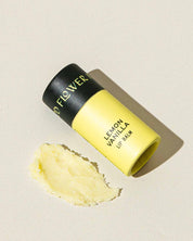 A yellow tube of Good Flower Farm’s TESTER — Lemon Vanilla Organic Lip Balm with a black cap rests on a light surface beside a small smear of the moisturizing balm.