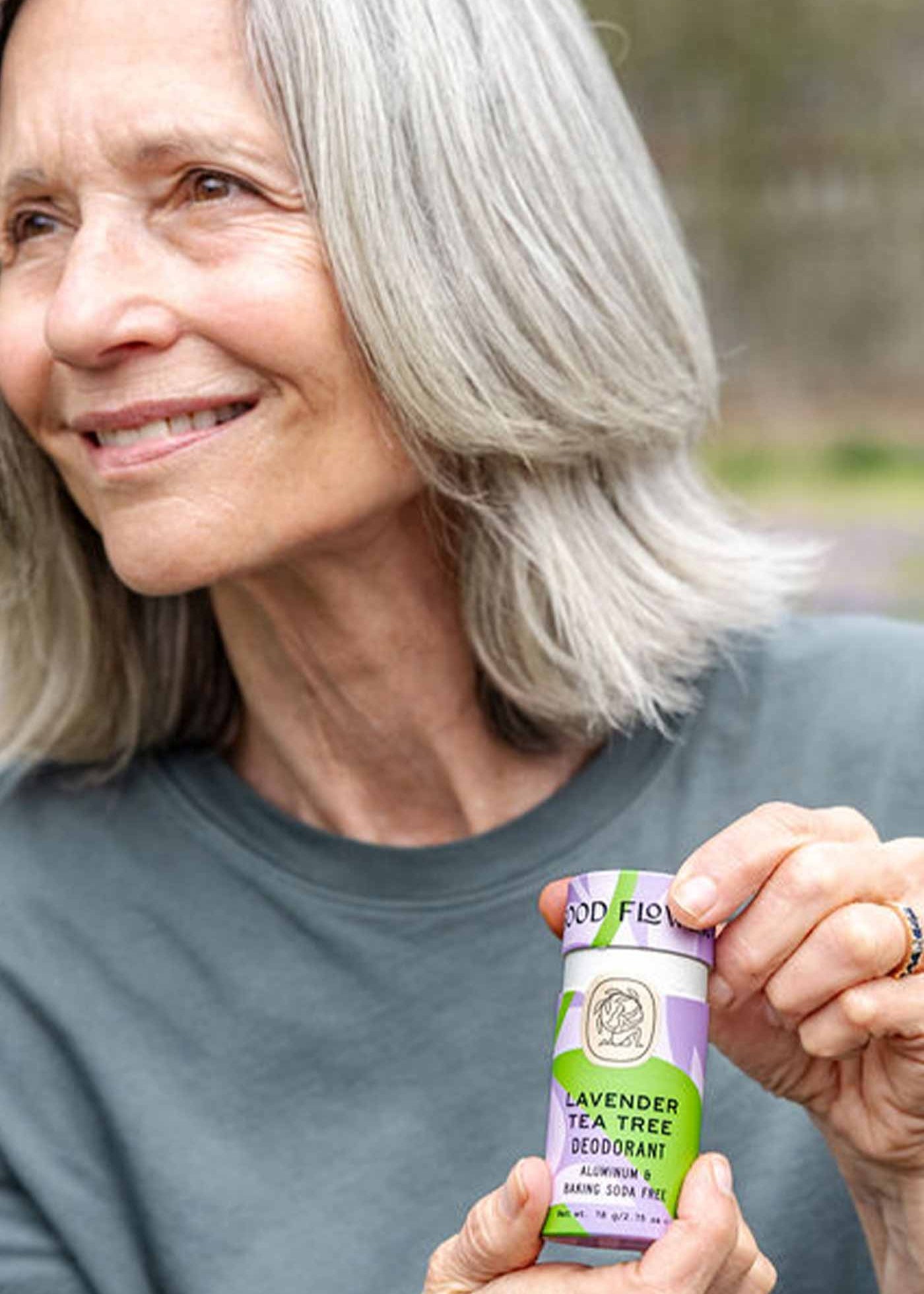 An older woman with gray hair smiles outdoors while holding a purple and green tube of Good Flower Farm Lavender Tea Tree Deodorant in a plastic-free tube.
