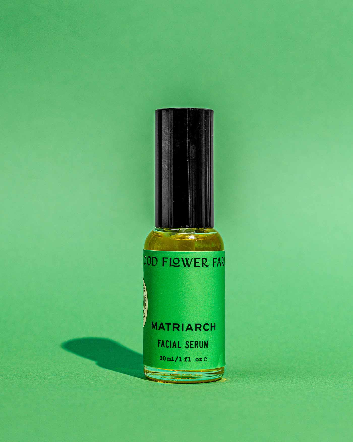 A small glass bottle of Good Flower Farm Matriarch Facial Serum, featuring a black cap and green label, stands upright on a green background. Infused with squalane, the serum casts a shadow on the surface.