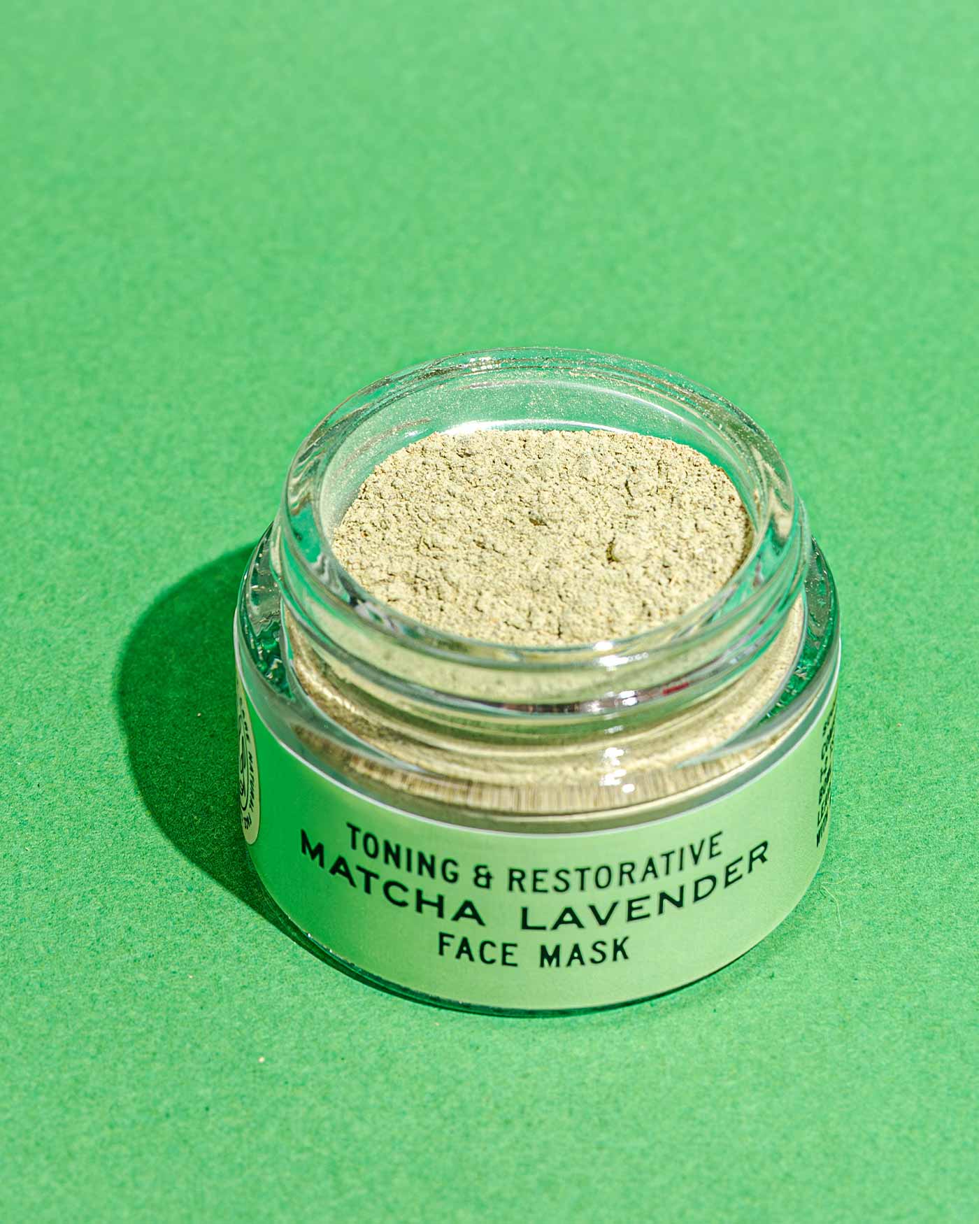 A small glass jar of Good Flower Farms Matcha Lavender Botanical Face Mask sits on a green surface, filled with pale green matcha powder and French green clay. The lid is off, displaying the soothing botanical blend inside.