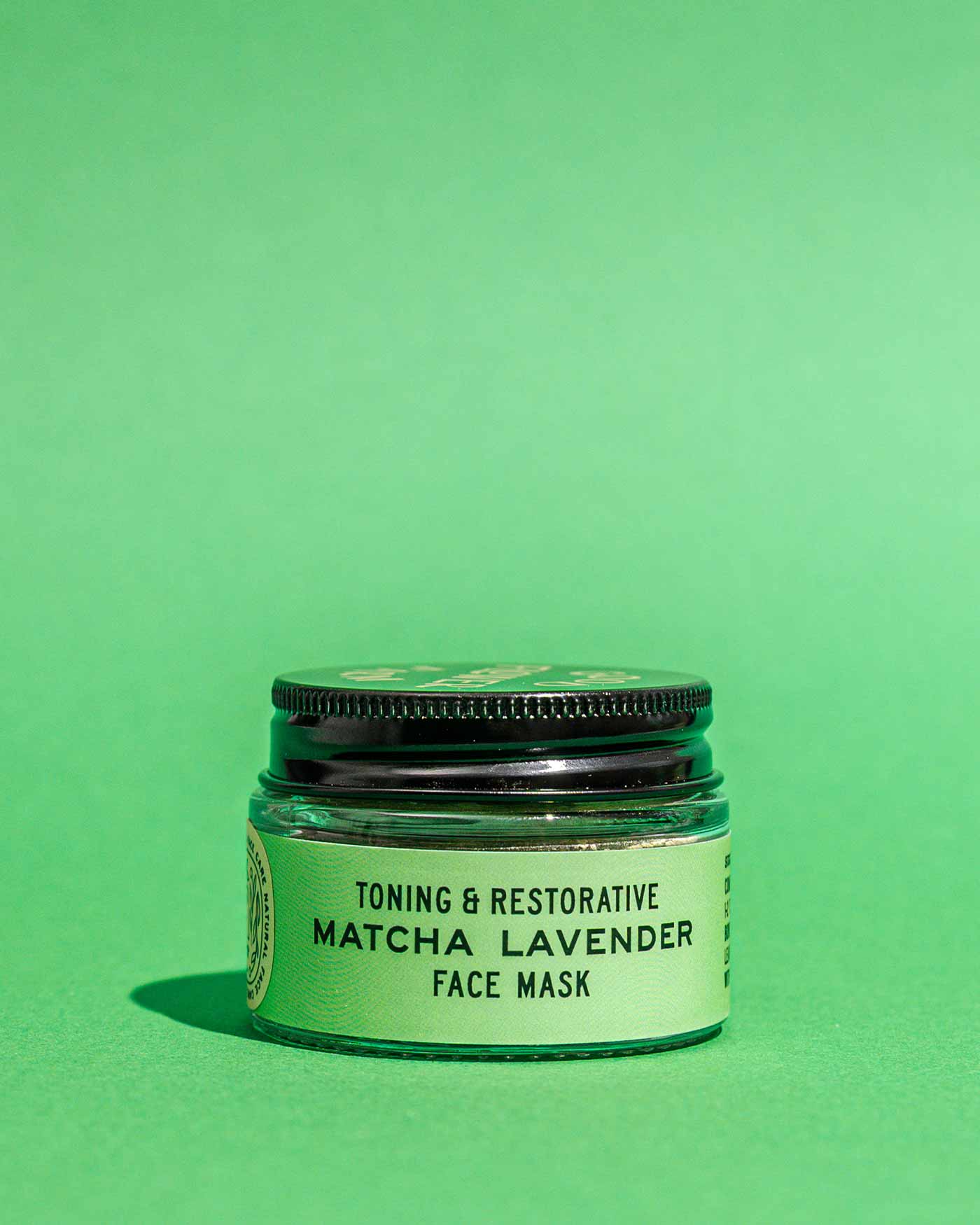 A small jar of Good Flower Farm’s Matcha Lavender Botanical Face Mask, featuring a green label and black lid, sits on a solid green background.