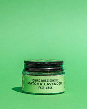 A small jar of Good Flower Farm’s Matcha Lavender Botanical Face Mask, featuring a green label and black lid, sits on a solid green background.